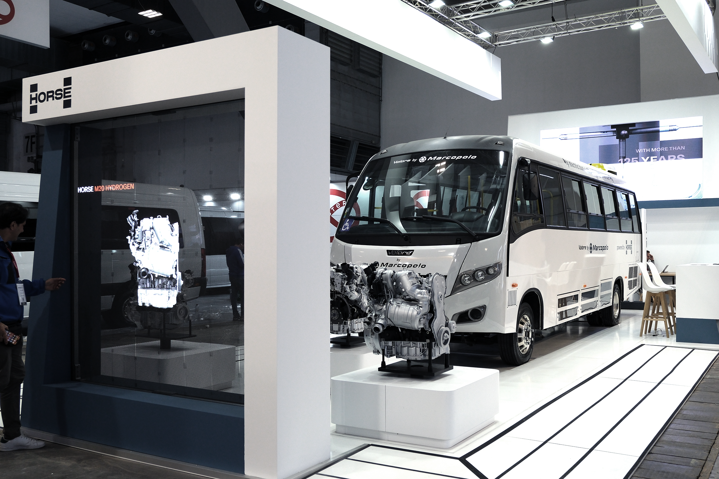 一辆印有 Marcopolo 和 Horse Powertrain 品牌的巴士停在活动展区的屏幕前和展示的发动机后面