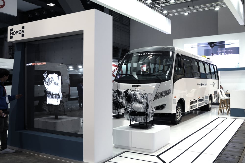 一辆印有 Marcopolo 和 Horse Powertrain 品牌的巴士停在活动展区的屏幕前和展示的发动机后面